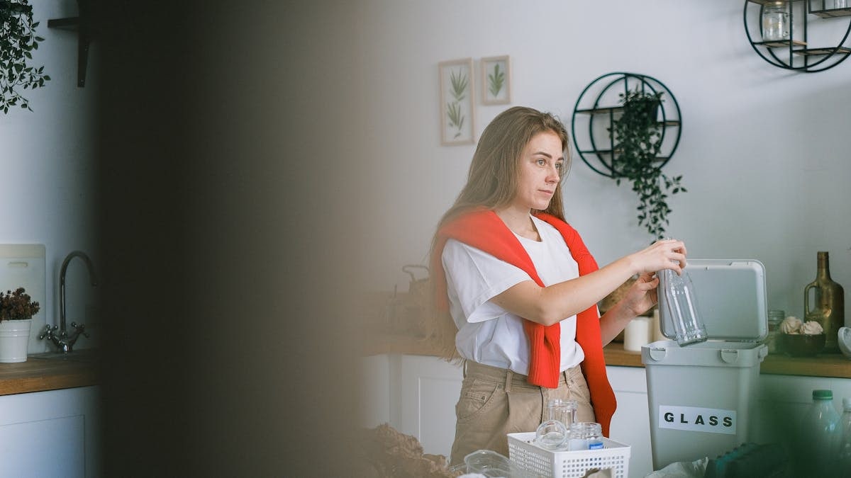 Young woman looking away while standing near table with rubbish in kitchen and collecting glass into special bucket
