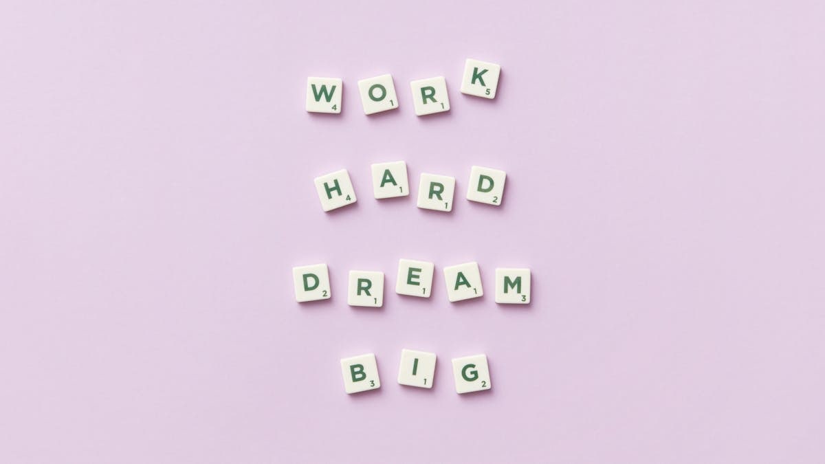 Scrabble tiles spell 'Work Hard Dream Big' on a pastel purple surface, top view.